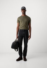 Olive green short-sleeve t-shirt, dark jeans, brown shoes, and black handbag. A gray cap with text rests on the head. Simple design, no patterns.