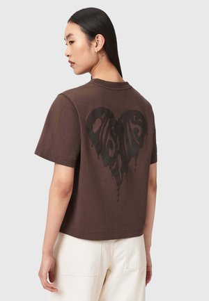 Kvinde med langt mørkt hår iført en brun t-shirt med et sort dryppende hjerte design på ryggen og hvide bukser.