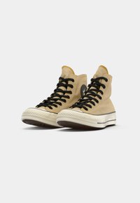 Baskets montantes en toile beige clair avec des lacets noirs, un logo rond noir et un embout et une semelle en caoutchouc blanc.