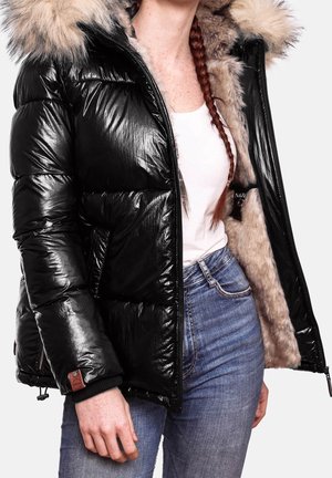 Personne portant une veste matelassée noire brillante avec doublure en fourrure et capuche, chemise blanche, jean bleu et cheveux rouges tressés sur l'épaule.
