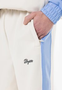 Hvide atletiske bukser med sidelomme, lyseblå accenter og broderet logo. Glat tekstur med elastisk talje.