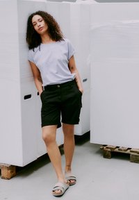 Ung kvinna med lockigt hår, iklädd en ljusgrå t-shirt, svarta shorts och sandaler, lutande mot inpackade pallar i en industrilokal.