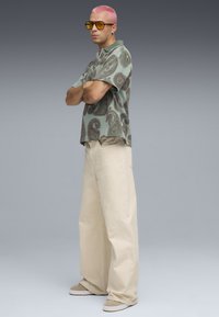 Short-sleeve, green paisley-patterned polo shirt, cream wide-leg pants, beige shoes, and yellow-tinted sunglasses. Smooth texture, relaxed fit.