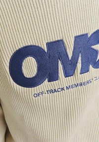 Jersey de punto crema con un gran logo bordado en azul marino de "OMK" y el texto "OFF-TRACK MEMBERS CLUB" debajo, también bordado a juego.
