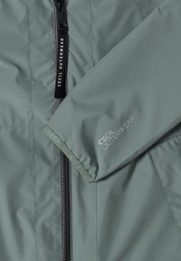 Hellgrüner Jackenärmel und Vorderteil mit schwarzem Reißverschluss, mit "CECIL OUTERWEAR" Logos auf dem Ärmel und am Reißverschlusszieher.