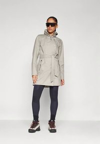 Sieviete, tērpusies pelēkā jostotā lietusmētelī, melnās legingās, pārgājušo zābaku un lielās melnās saulesbrillēs, stāv pretī baltai fona.