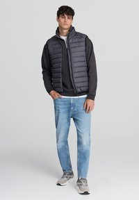 Szara kamizelka puchowa założona na czarną bluzę, w połączeniu z jasnoniebieskimi dżinsami i szarymi trampkami. Kurtka ma wysoki kołnierz i zamek błyskawiczny.