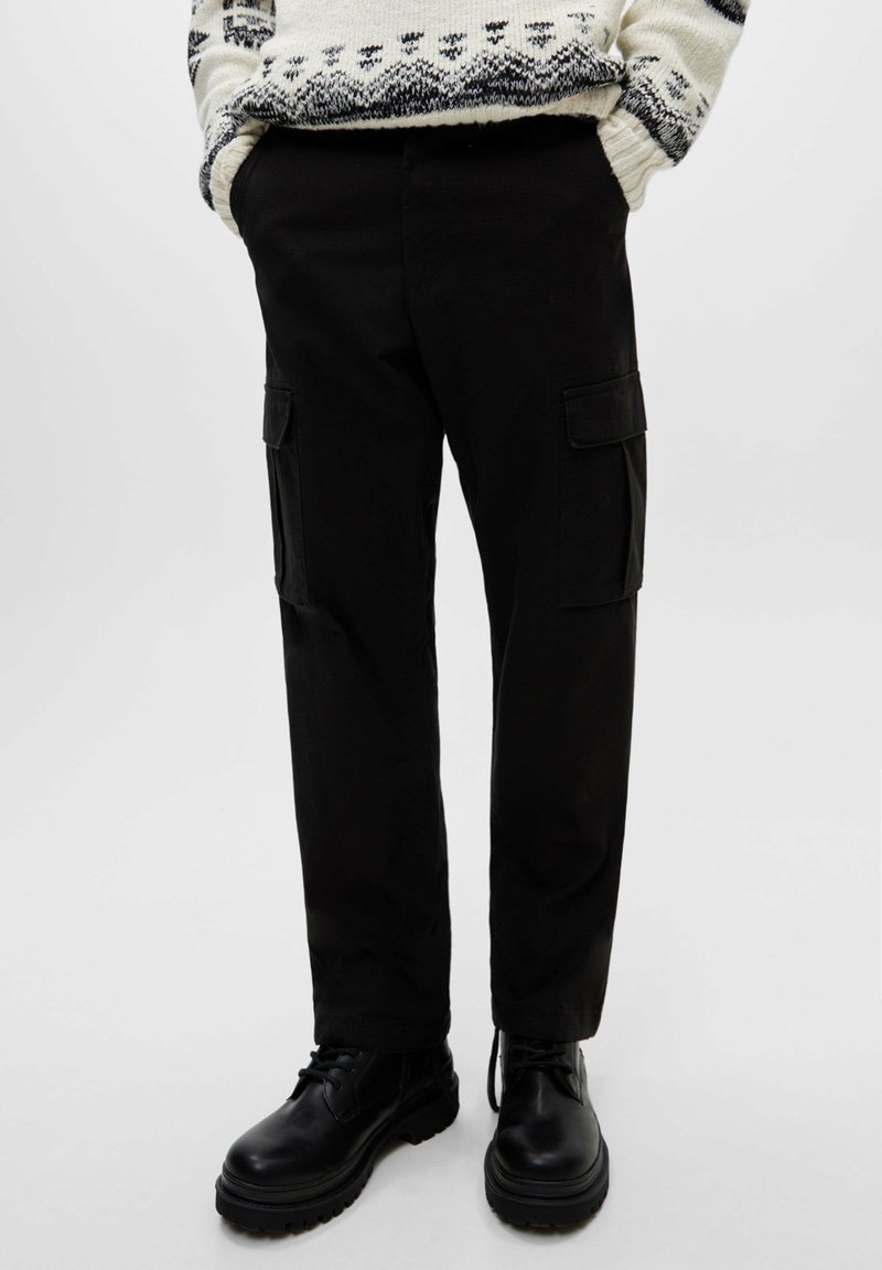 Osoba v černých cargo kalhotách s bočními kapsami a černých šněrovacích botách, se rukama v kapsách, stojící na světlém jednobarevném pozadí.