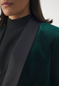 Grön sammetsblazer med svart satin sjalkrage, med en mjuk textur och skräddarsydd passform, delvis visad mot en neutral bakgrund.