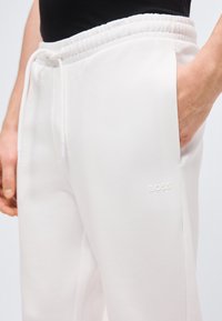 Λευκή φόρμα με ελαστική μέση με κορδόνι, πλαϊνές τσέπες και ανάγλυφο λογότυπο "BOSS" στο αριστερό μηρό, φορεμένη με μαύρο τοπ.