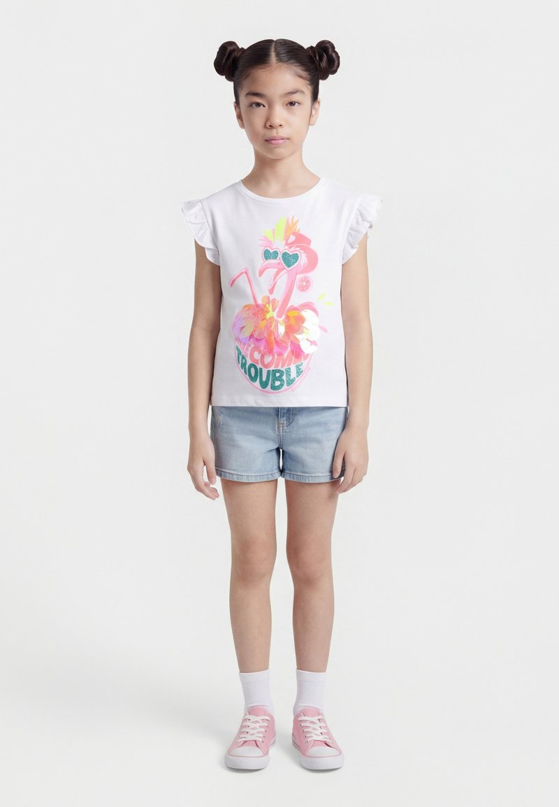 Κορίτσι με τα μαλλιά σε δύο κότσους, που φορά λευκό αμάνικο τοπ με βολάν και πολύχρωμο μοτίβο φλαμένγκο, τζιν σορτς, λευκές κάλτσες και ροζ αθλητικά παπούτσια.