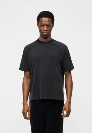 Jeune homme debout, de face, portant un T-shirt noir uni à manches courtes et un pantalon noir, sur un fond blanc.