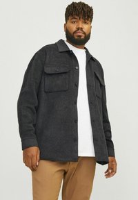 Dark gray flannel shirt with a collar, two chest pockets, and button closures, worn over a white t-shirt and paired with light brown pants.