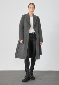 Palton gri din lână cu guler ascuțit, închidere cu un singur nasture și două fante laterale, asortat cu pantaloni din piele negri și cizme cu șireturi.