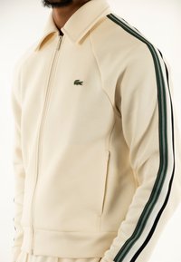 Chaqueta cremosa con cremallera y cuello puntiagudo, con acentos a rayas verdes y negras a lo largo de las mangas y un pequeño logo bordado.