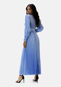 Abito lungo azzurro chiaro plissettato con maniche lunghe a sbuffo, corpetto aderente e fondo fluido. Abbinato a sandali neri con tacco alto.