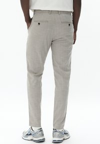Beige Stoffhose mit strukturiertem Muster, zwei Gesäßtaschen und schlankem Schnitt. Kombiniert mit grauen Sportschuhen.