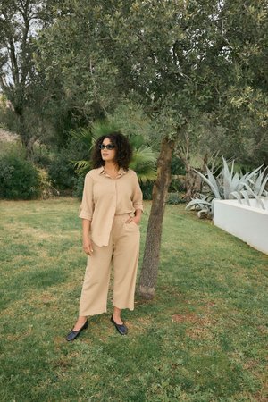 Femme en tenue beige et chaussures noires, debout, une main dans la poche, sous un arbre feuillu dans un jardin herbeux.