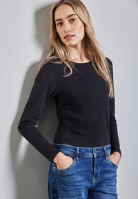 Musta pitkähihainen toppi pehmeästä kankaasta yhdistettynä keskisinisiin farkkuihin. Istuvuus on rento, pyöreä kaula-auko ja etutaskut.