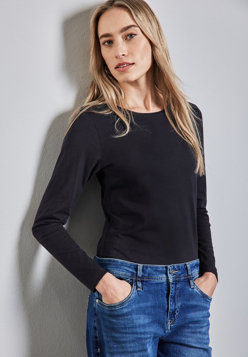 Musta pitkähihainen toppi pehmeästä kankaasta yhdistettynä keskisinisiin farkkuihin. Istuvuus on rento, pyöreä kaula-auko ja etutaskut.