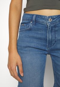 Acercamiento de una persona que lleva jeans azules y una blusa corta oscura, mostrando la cintura, la cadera y la mano izquierda descansando junto al muslo.