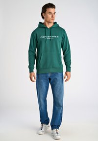 Felpa verde con tasca a canguro frontale e testo bianco "COPENAGHEN". Indossata con jeans blu e sneakers bianche. Tessuto liscio, vestibilità casual.