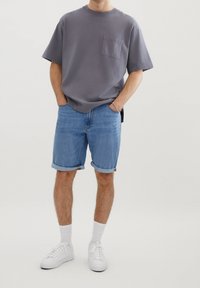 Camiseta gris de manga corta con un bolsillo en el pecho, combinada con pantalones cortos de denim azul claro con dobladillos enrollados. Terminada con zapatillas blancas y calcetines.