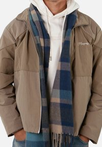 Giacca zip-up marrone sopra una felpa bianca con cappuccio, abbinata a una sciarpa a quadri blu con accenti marrone chiaro. Visibile una notevole texture e stratificazione.