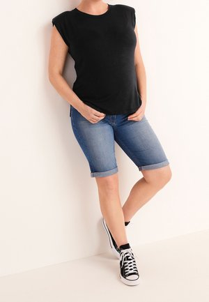 Kvinde iført sort ærmeløs top, blå op rullet denimshorts og sort-hvide sneakers står op ad en hvid væg.
