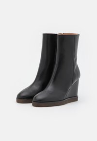 Botas de tobillo de cuña de cuero negro con un acabado suave, punteras redondeadas y una suela marrón texturizada. El diseño presenta una silueta elegante y minimalista.