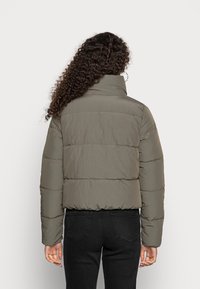 Oliwkowo-zielona pikowana kurtka puchowa z wysokim kołnierzem, pikowanym wzorem i elastycznymi mankietami. Noszona na czarnych jeansach, widoczna od tyłu.