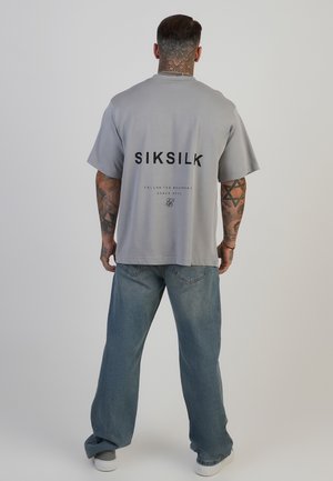 Tricou gri supradimensionat cu logo-ul negru "SIKSILK" pe spate, mâneci scurte și decolteu rotund; purtat cu blugi de culoare albastră deschisă și pantofi albi.