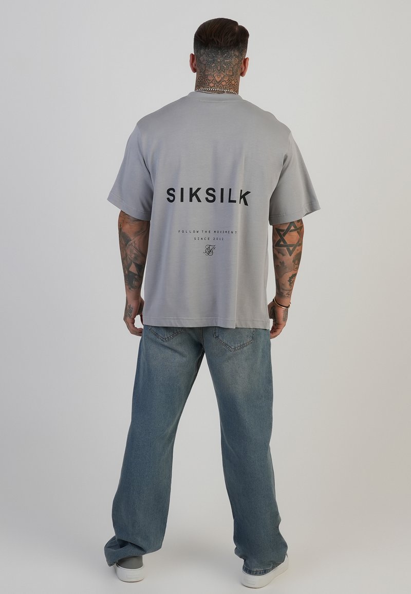 Hall hõlgatud t-särk musta "SIKSILK" logoga seljal, lühikeste varrukatega ja ringsuuga; kantud helesiniste teksade ja valgete kingadega.