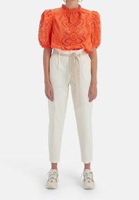 Oranje blouse met pofmouwen en oogjespatroon, gecombineerd met crèmekleurige tapering broek en beige sneakers, met een gestrikt ceintuur in de taille.