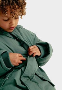 Zelená polstrovaná bunda s kapucí, má hladký vnější povrch, elastické manžety a přední zapínání. Kudrnaté vlasy a ruce, které upravují bundu.