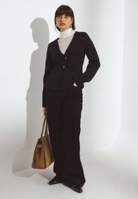 Must mustrit, millel on kaks nuppu, valge ujumispusa peal, mustad püksid, pruun suede kott käes ja jalas terava ninasügavustega mustad kingad.