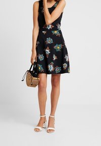 Žena v černých květovaných šatech bez rukávů, držící dřevěnou kabelku, v bílých sandálech na kotníku, stojící na jednobarevném pozadí.