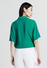 Blusa verde a maniche corte con colletto, realizzata in tessuto leggero e dalla vestibilità ampia, abbinata a pantaloni bianchi con dettagli in pizzo.
