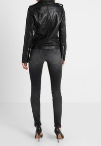 Kvinde iført en tætsiddende sort læderjakke, mørkegrå skinny jeans og sorte højhælede sko, stående foran en hvid baggrund.