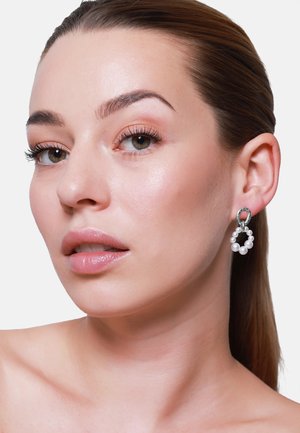 Silberne Creolen mit kleinen weißen Perlen, die ein strukturiertes Design aufweisen. Die Ohrringe hängen und ergänzen einen natürlichen Makeup-Look.
