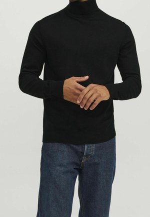 Homme portant un pull à col roulé noir et un jean bleu, les mains jointes devant, sur un fond clair uni.