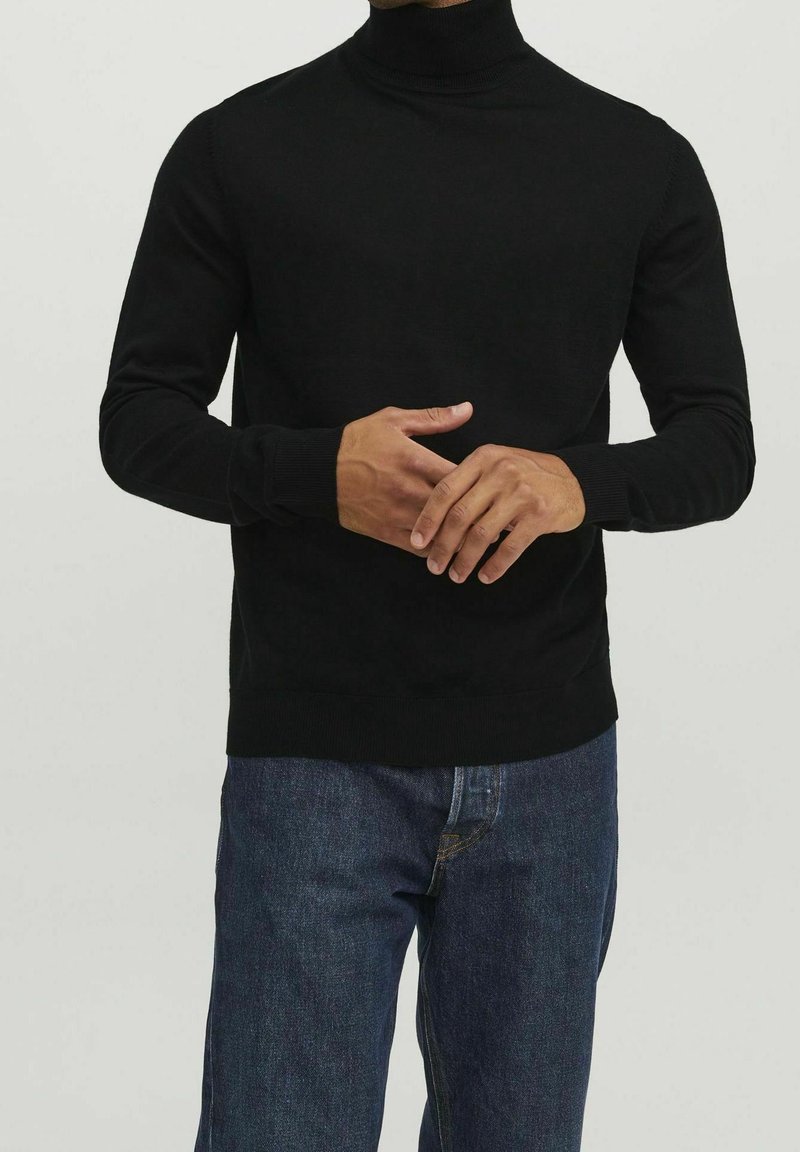 Homme portant un pull à col roulé noir et un jean bleu, les mains jointes devant, sur un fond clair uni.