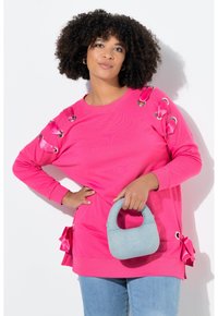 Roze sweatshirt met oogjesaccenten en satijnen linten op de mouwen. Lichtblauwe handtas met een gebogen handvat. Het model combineerde dit met denim onderstukken.