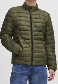 Olivgrüne Steppjacke mit hohem Kragen, horizontalem Steppmuster, Reißverschluss vorne und kleinem Markenlogo auf der Brust.