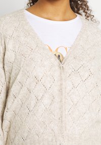 Zbliżenie na osobę noszącą kremowy sweter w romby na biały podkoszulek z częściowym widocznym wzorem graficznym.