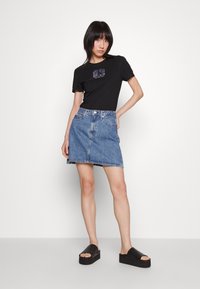 Camiseta de algodón negra con diseño impreso, combinada con una falda de mezclilla azul claro. El modelo lleva sandalias de plataforma negras. Fondo simple.