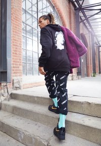 Personne avec des dreadlocks montant des escaliers extérieurs en béton, portant un sweat à capuche noir avec un visage blanc imprimé, un pantalon noir avec le texte "OBEY" et tenant une veste violette.