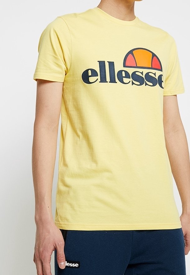 Personne portant un t-shirt à manches courtes jaune avec un grand logo "ellesse" noir et un demi-cercle orange-rouge au-dessus du texte.