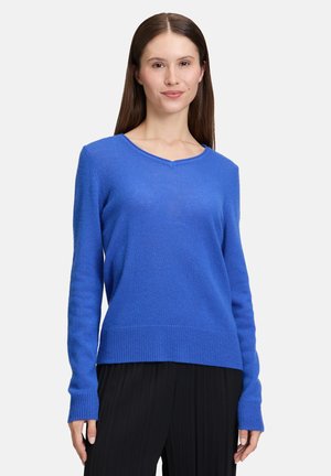 Woman with straight brown hair wearing a bright blue knit sweater and black pleated pants against a white background.