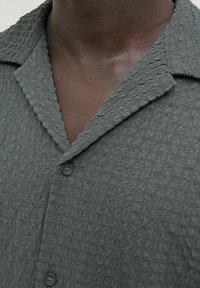 Camisa texturizada verde oscura con cuello con muesca, dos botones y una tela con un patrón sutil que presenta un efecto arrugado.